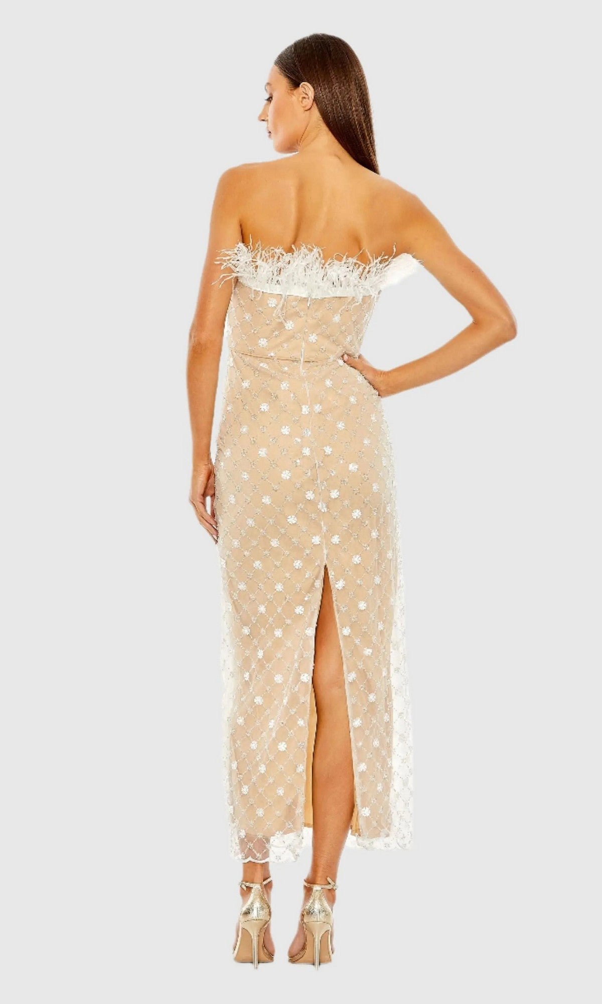 A woman stands with her back to the camera in gold high-heeled sandals, wearing the Mac Duggal 68140V Tea-Length Party Dress—a stunning strapless design featuring embellished sheer beige fabric, white floral patterns, and elegant feather trim.